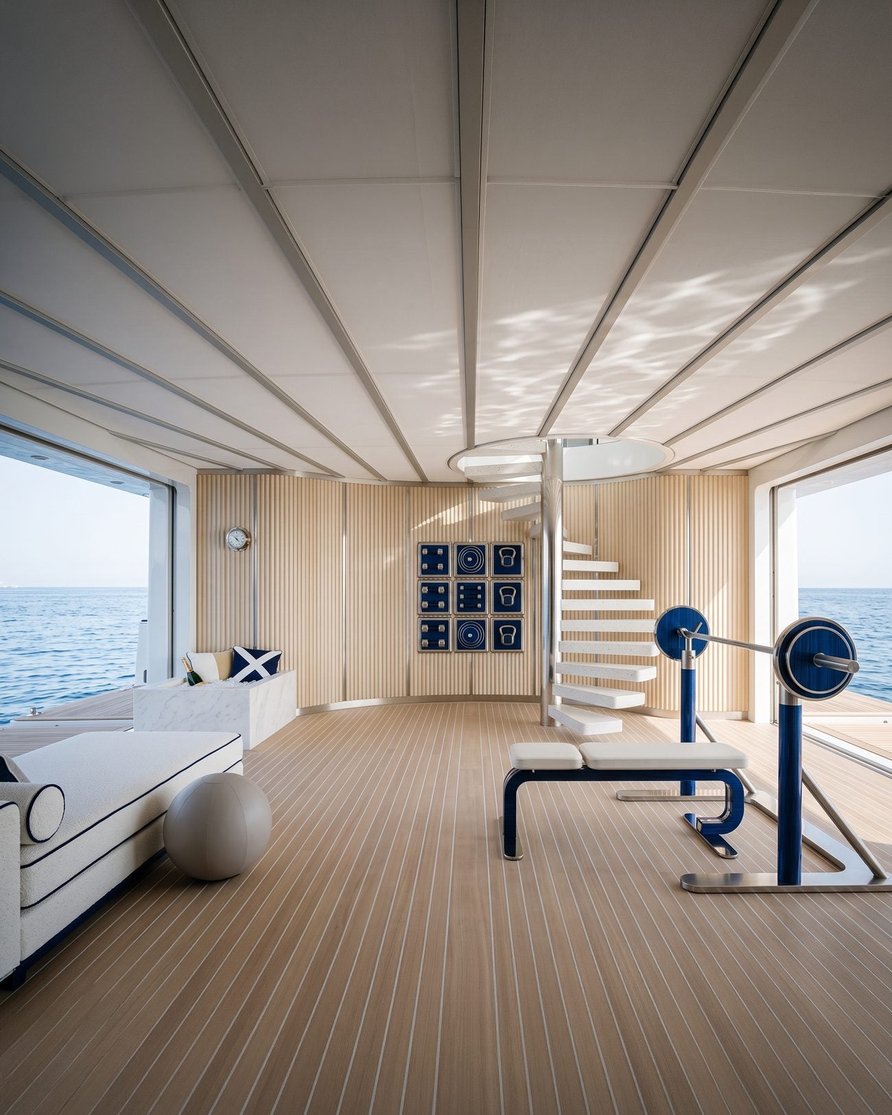 A superyacht gym in pale wood and navy blue, with an Alama bench, barbell and framed wall display, open to the sea.