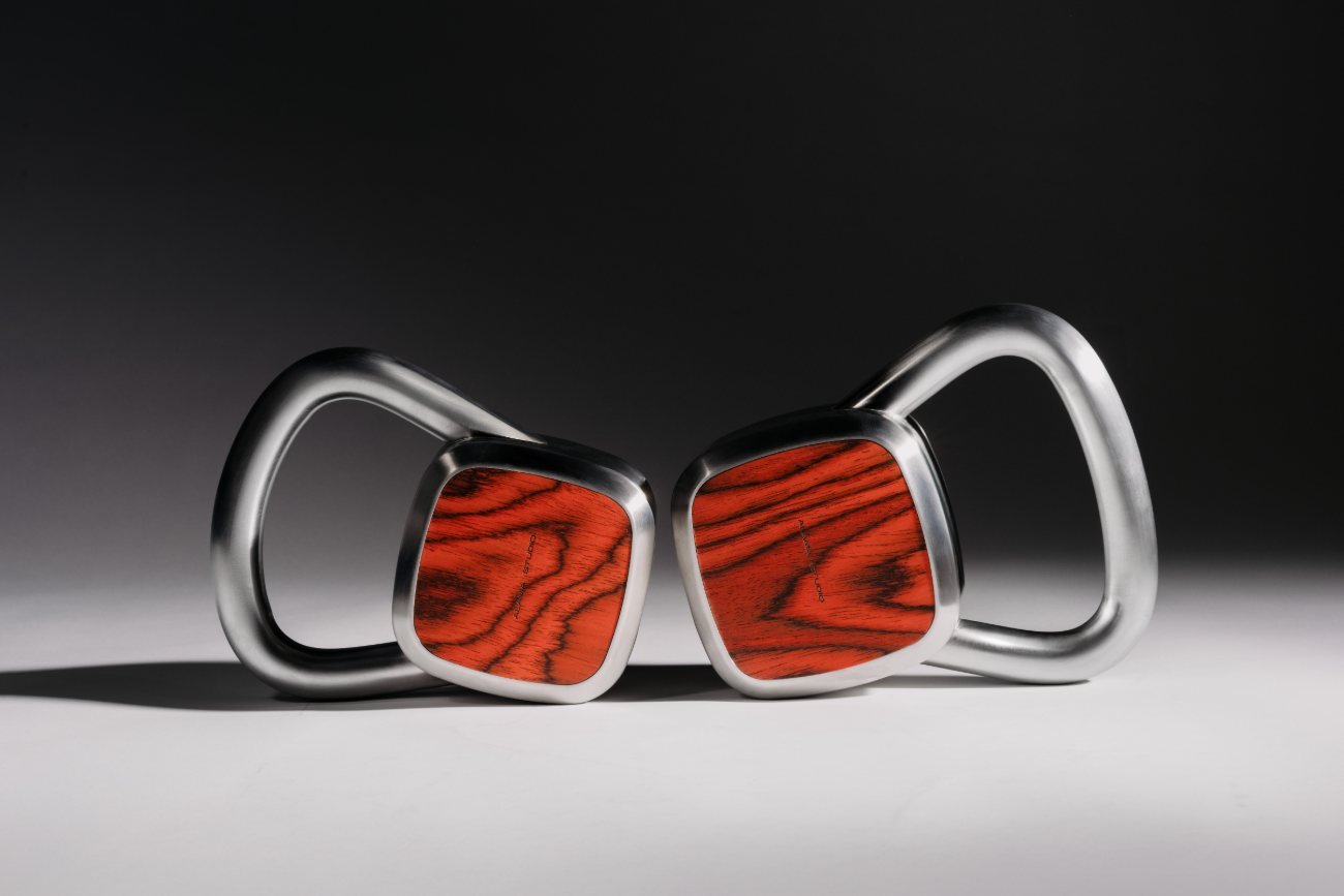 A sculptural pair of Alama weights in brushed steel and red wood, lit like design objects within a designed interior rather than on a gym floor.