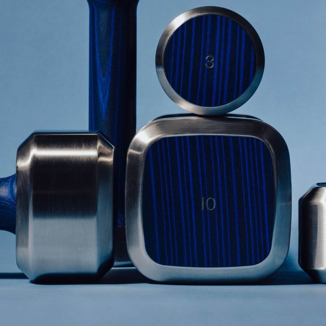 A compact, coordinated set of small Alama weights: a round and a square plate engraved 3 and 10 in steel and blue ALPI wood, beside a short dumbbell.