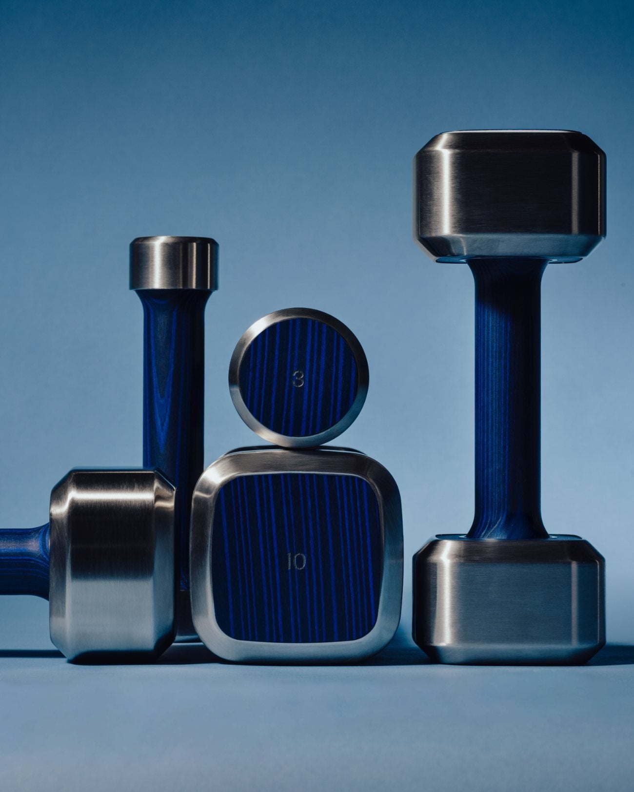 An Alama freeweights set in steel and deep-blue wood, photographed against a marine blue backdrop.