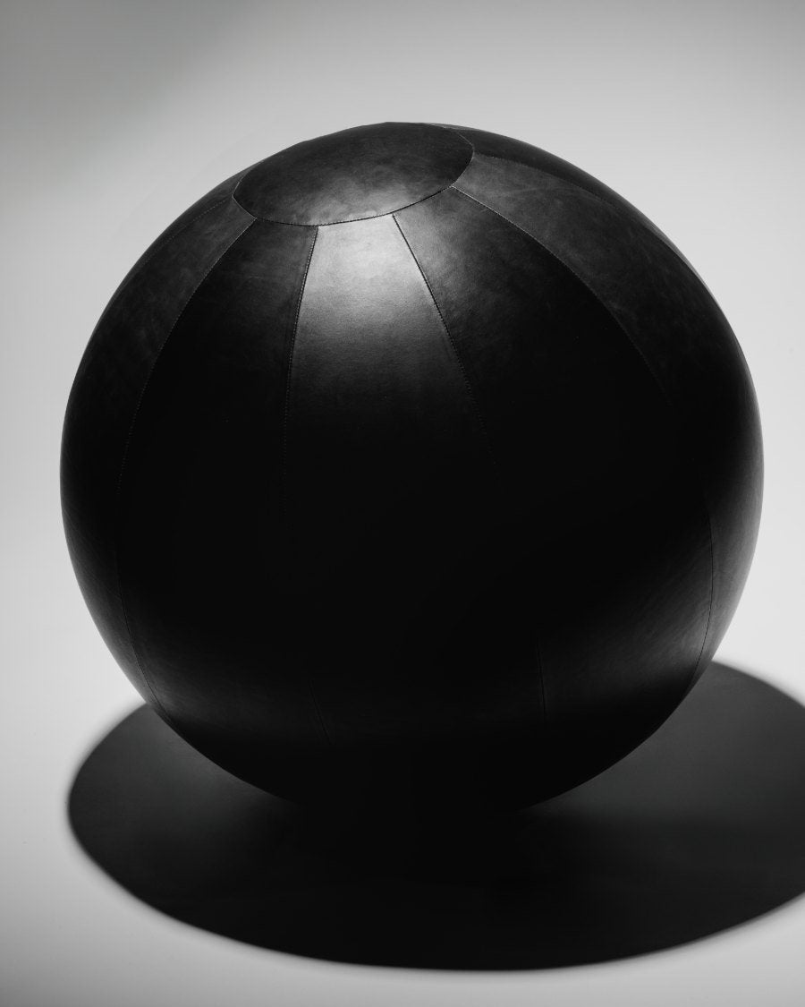 An Alama yoga ball in panelled black leather, lit as a sculptural object on a pale ground.